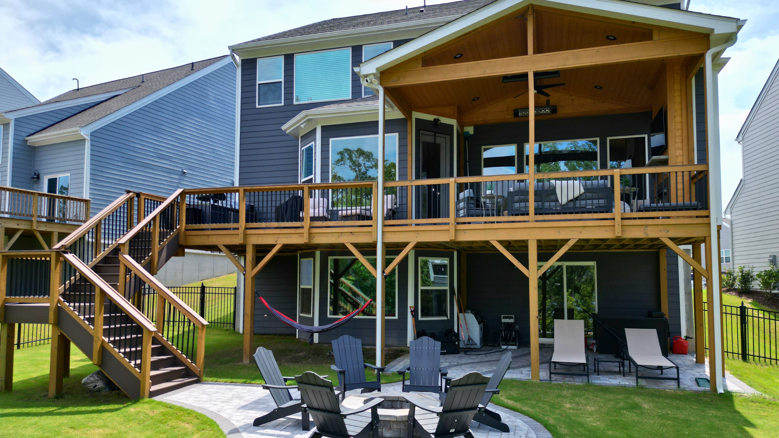 A two-story backyard living space featuring an elevated wooden deck with a covered porch section and black railings. Below the deck is a stone patio with a fire pit surrounded by Adirondack chairs, a hammock, and a lounge area under the deck's structure.