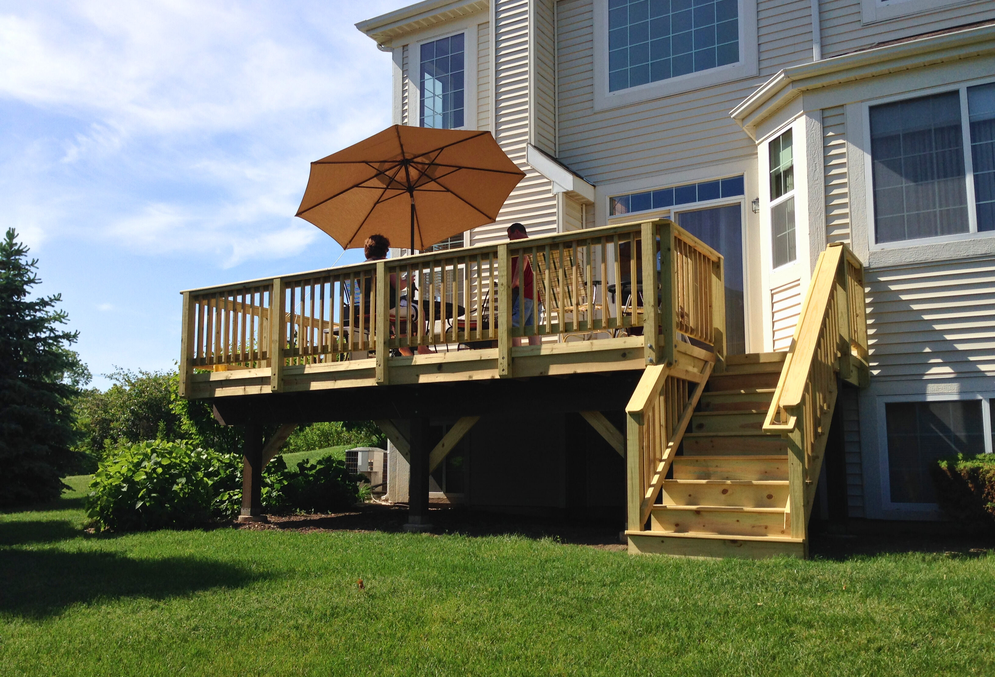 : A newly constructed elevated wooden deck with natural wood railings and steps, featuring an umbrella and outdoor furniture, attached to a light-colored house.