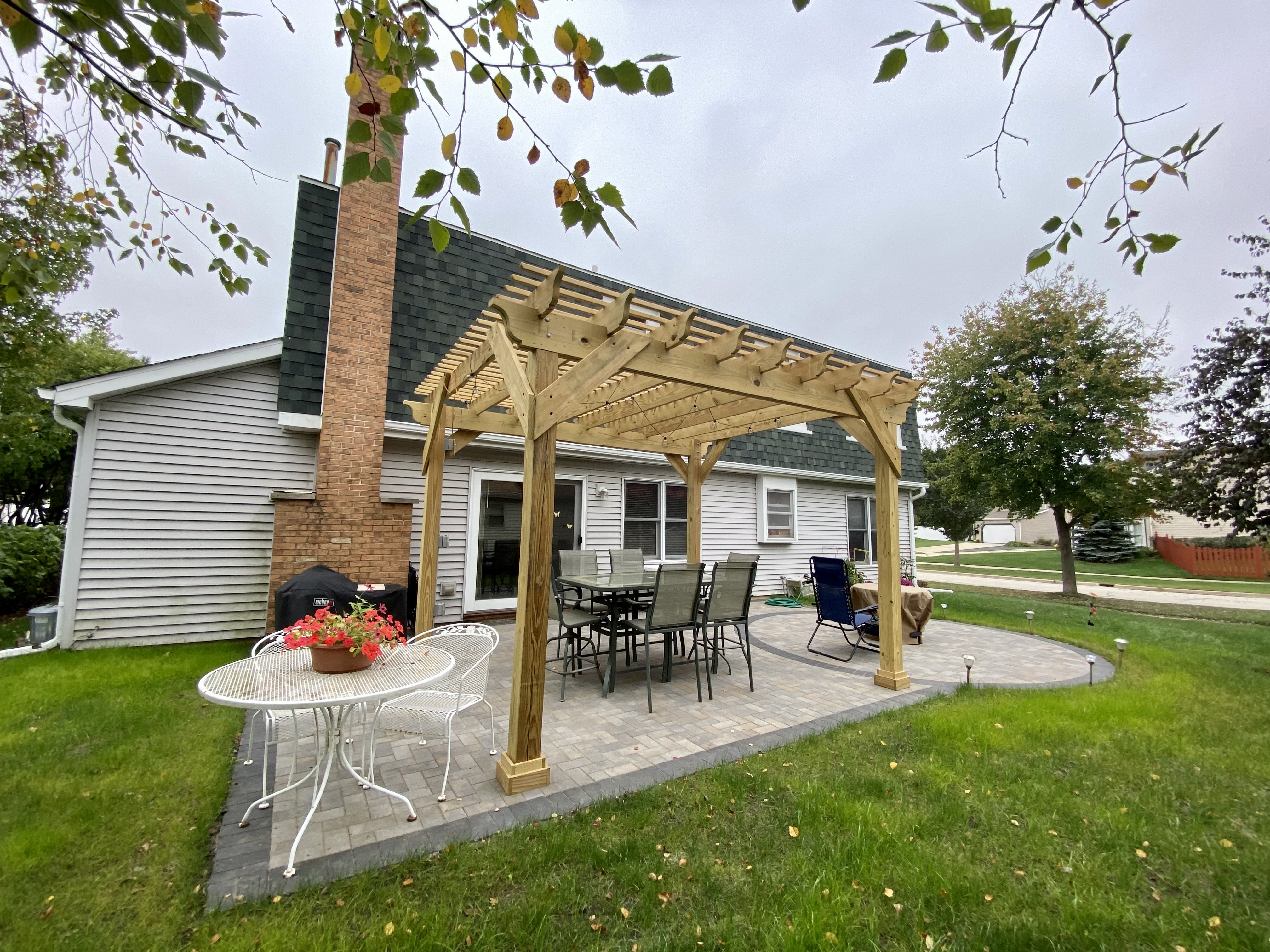 Wood pergola on a paver patio with dining furniture surrounded by grass. 