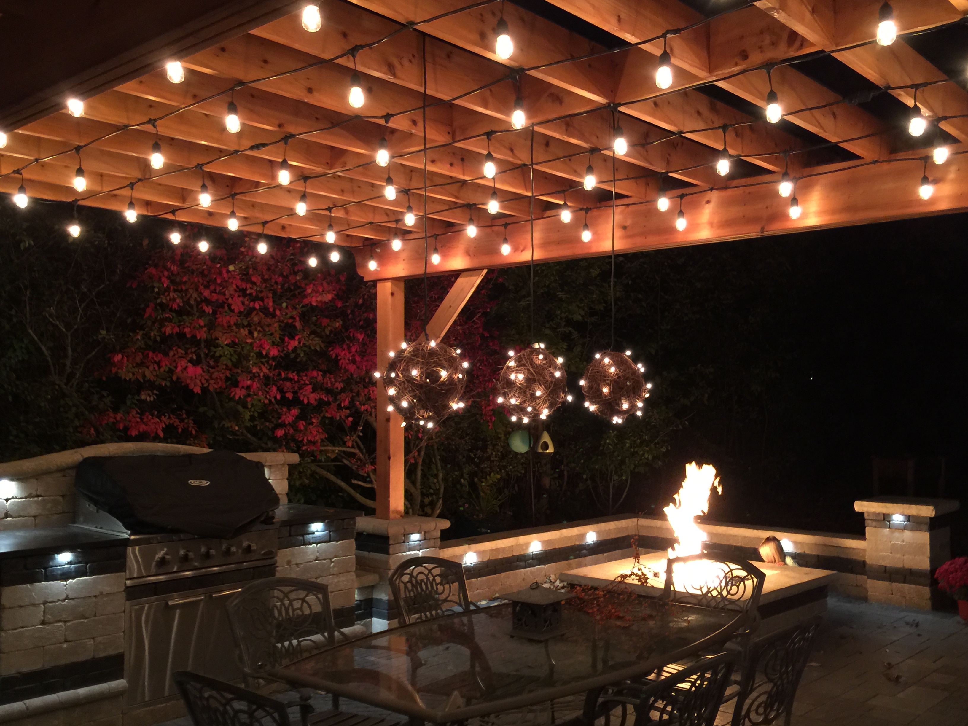 Custom pergola, outdoor kitchen, and fire pit at night with string lights.