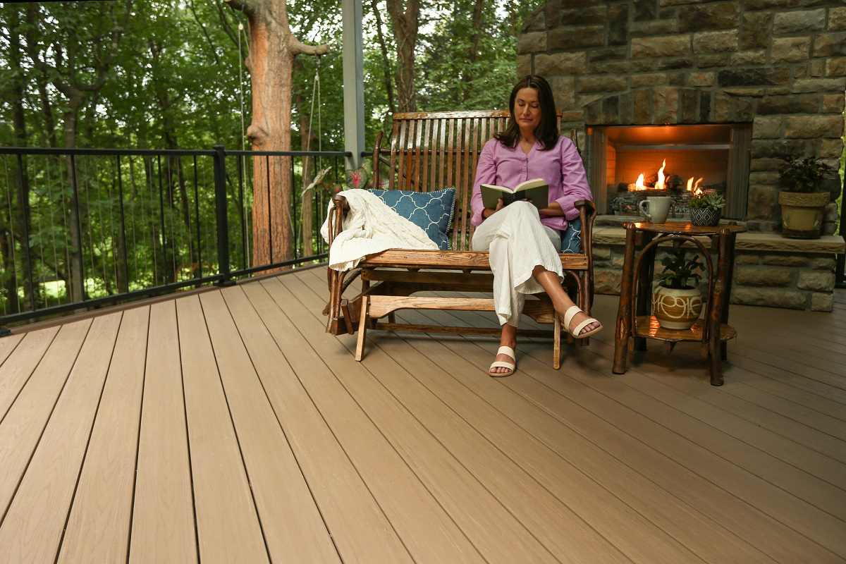 Person reading in front of stone fireplace and sitting on a wood rocking bench that is located on the tan deck with a black railing