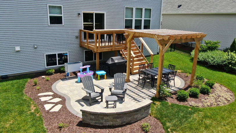 An elevated modern composite deck with light grey planks, dark grey and wood-toned railings, and integrated lighting, featuring contemporary outdoor seating, attached to a dark grey-sided home with large windows and sliding glass doors in Geneva, IL. A retractable screen is visible on the left side of the deck.