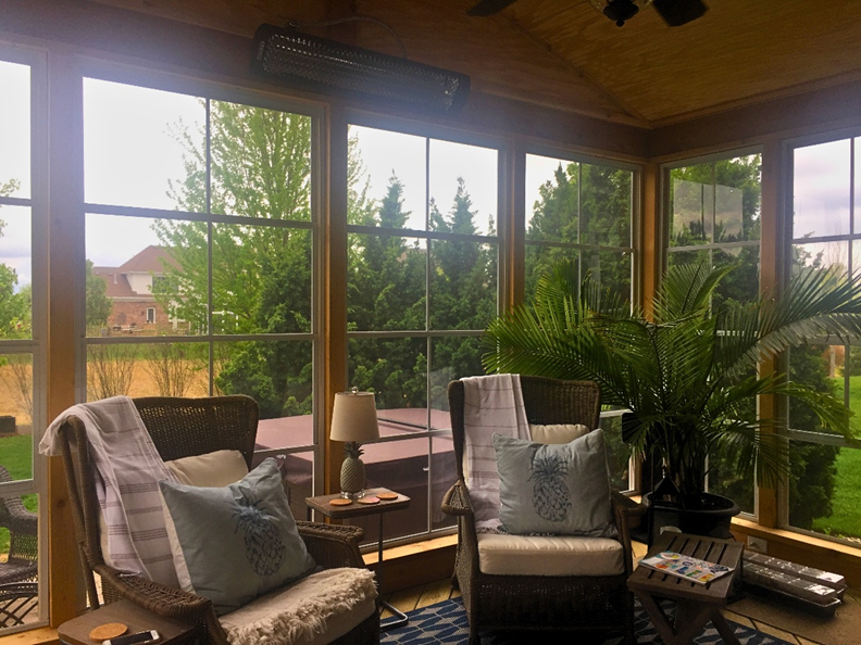 The interior of a bright and inviting screened-in porch with light-colored wood paneling, multiple large windows, comfortable wicker furniture with blue and white cushions, a ceiling fan, and a view of green trees and a hot tub outside, in Geneva, IL.