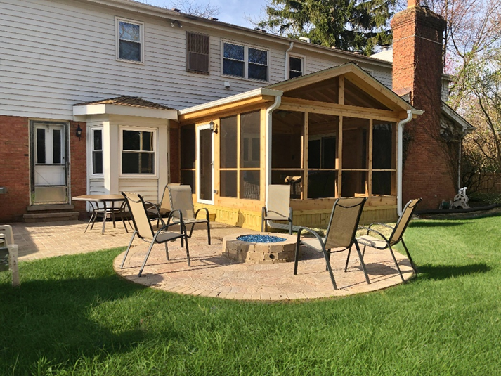 A backyard patio with a circular fire pit area and a newly built screened porch attached to a brick and siding house, surrounded by green grass.