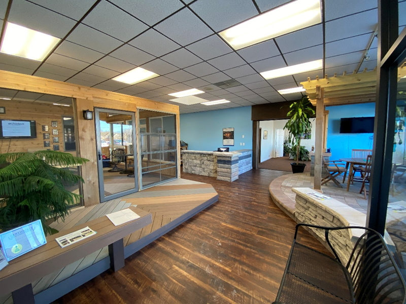 The interior of a modern showroom featuring various outdoor living displays. There are sections with wood and composite decking, stone accents, glass patio doors, and seating areas, showcasing different design possibilities.