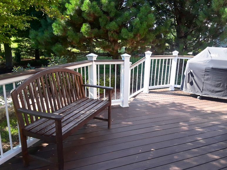  A dark brown composite deck with white railings and balusters, featuring a wooden bench and a covered grill, surrounded by lush green trees and foliage, on a sunny day in Geneva, IL.