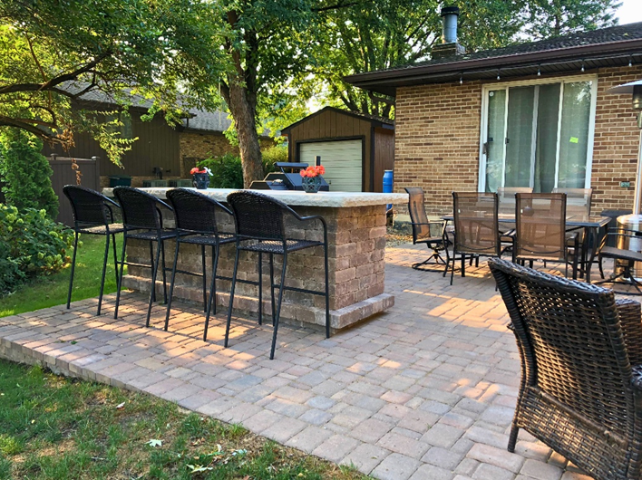 Spacious paver patio with a brick outdoor bar and black bar stools, surrounded by seating areas and mature trees in a Homer Glen backyard.