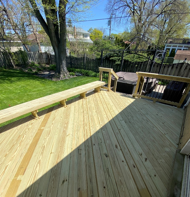 A new, light-colored wooden deck with boards running lengthwise. A long, matching wooden bench is integrated into the deck's railing on the left. In the back right corner, a dark spa is visible, enclosed by the deck railing. A green lawn and trees are in the background.