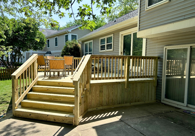 A large, light-colored stone paver patio extends from the back of a beige-sided house with white French doors. A small brown deck with steps connects the doors to the patio. Outdoor furniture and an umbrella are set up on the left side of the patio. 
