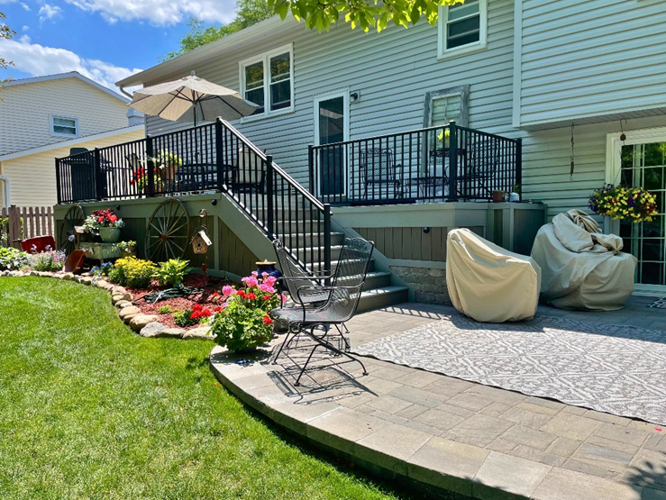 A multi-level composite deck with black railings on the back of a light gray house. Stairs lead down to a stone paver patio. A decorative garden with colorful flowers and rustic wagon wheels is adjacent to the deck.