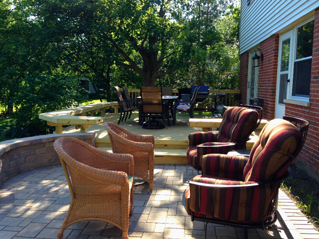 A multi-level outdoor living space featuring a brick patio with various seating options and a wooden deck with a dining set, surrounded by lush trees.