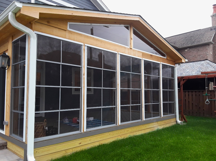 A new, three-season porch addition with a light wood-framed structure and large floor-to-ceiling windows. The base of the porch is a light-colored wooden deck. A white downspout runs down the right side of the structure.