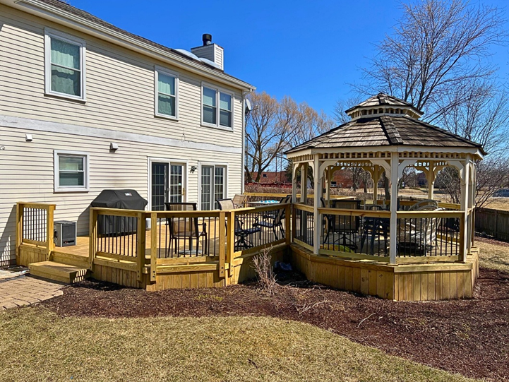 An aerial view of a large, multi-level natural wood deck attached to the back of a light-colored house. The deck has various seating areas, including a dining table, a lounge set, and benches.