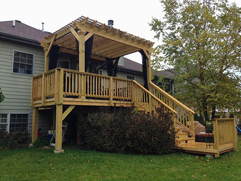 An elevated natural wood deck with a matching pergola and black privacy screens, featuring stairs leading down to a small landing, attached to a light green-sided house in Geneva, IL, with shrubs underneath.
