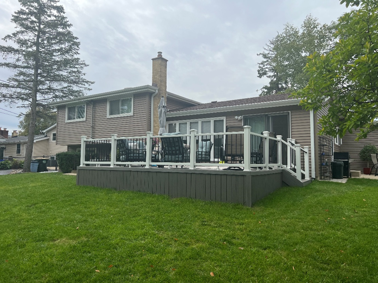 A backyard view of a light brown house with a large, ground-level composite deck. The deck has gray vertical siding around the base and a white railing with black metal balusters. Outdoor furniture is arranged on the deck.
