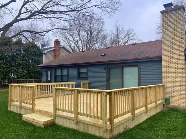 A newly built wooden deck with natural wood railings and steps, attached to the back of a house with blue siding and a brick chimney, overlooking a green lawn.