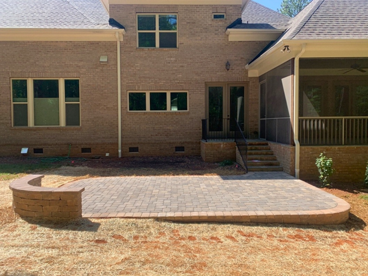 A newly installed stone paver patio with a curved brick retaining wall, leading up to double French doors on a brick home in Mint Hill, NC, with dry grass around it.
