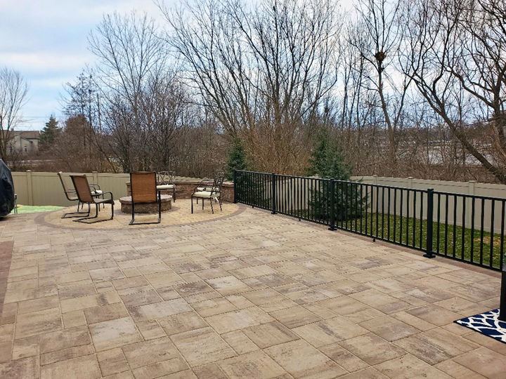 A large, multi-toned stone paver patio with a curved edge and a built-in stone fire pit surrounded by chairs. A black metal railing separates the patio from a grassy area and a white fence in the background.