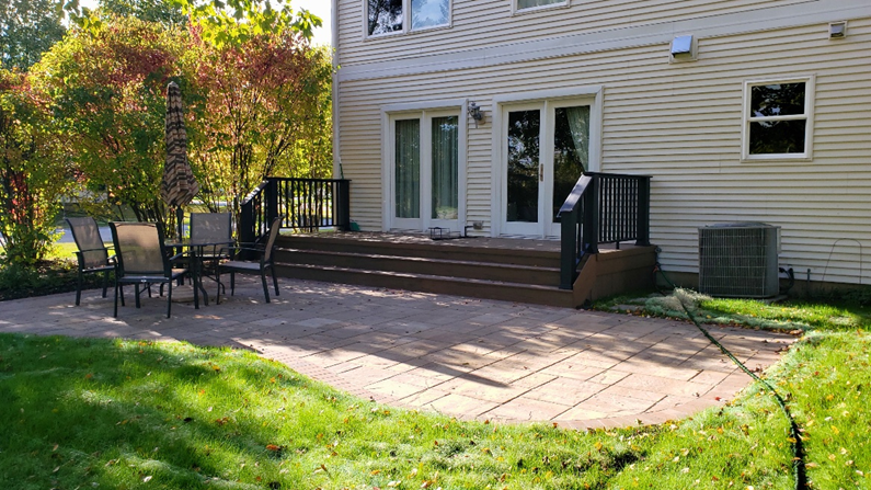 A large natural wood deck attached to a light beige house. The deck extends around to a multi-sided gazebo with a shingled roof, both featuring black railings. Outdoor furniture and a grill are visible on the deck.