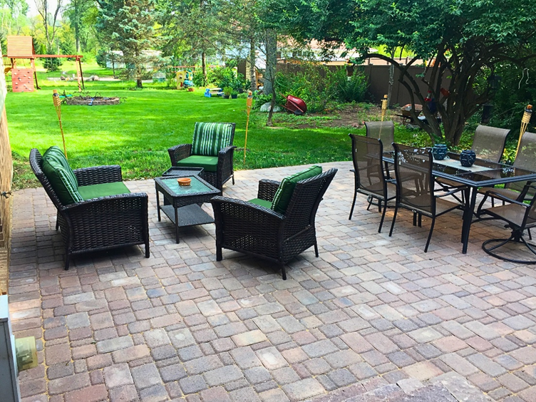 Brick paver patio with green wicker chairs, a coffee table, and a dining table with chairs, surrounded by lush greenery in Homer Glen.
