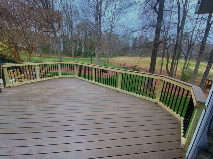 A wide, curved composite deck with brown planks and a light wood railing with black balusters, overlooking a grassy yard and wooded area under an overcast sky in Mint Hill, NC