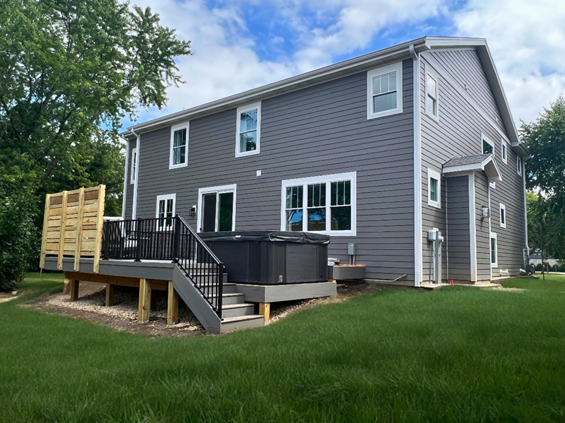 A two-story modern house with dark gray siding and white-framed windows. A new deck with composite decking and dark railings is attached to the back of the house. A hot tub is partially recessed into the deck, and a custom wooden privacy screen is visible on the left side.