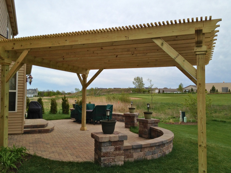 A spacious stone paver patio with a curved seating wall, covered by a large wooden pergola, leading to a home with a grill visible on the left, set in a wide-open grassy backyard in Geneva, IL.