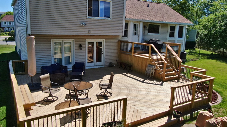 A newly constructed natural wood deck with a wide set of stairs leading down to a concrete patio. The deck features a dining set with wooden chairs and a table. It's attached to a light gray house with sliding glass doors.