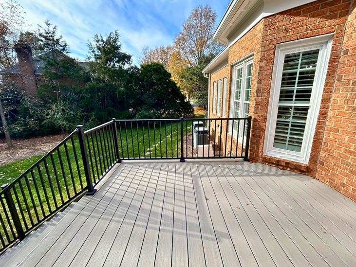 A modern grey composite deck with black metal railings and stairs, attached to a traditional brick home with white windows, overlooking a green lawn in Mint Hill, NC.