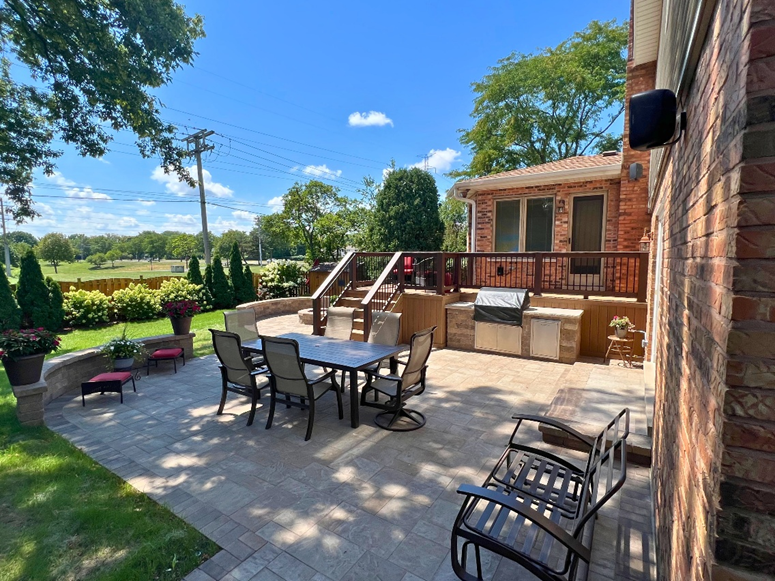 spacious backyard featuring a light-colored paver patio with a dining table and chairs. A raised deck with dark brown railings and an outdoor kitchen area with a built-in grill is visible on the right. A large green lawn is in the background.