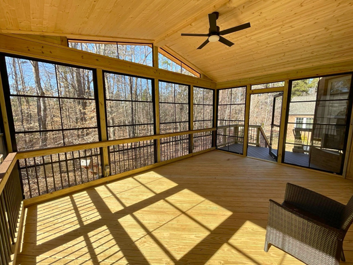 The bright interior of a newly constructed three-season room with light wood walls and ceiling, a ceiling fan, and large windows offering panoramic views of a wooded area in Mint Hill, NC