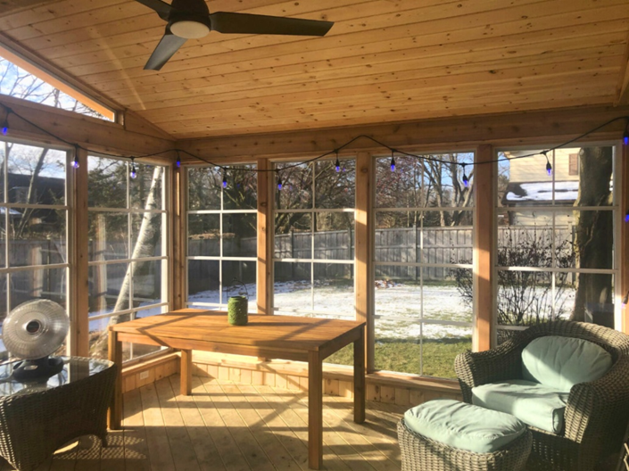 The interior of a three-season room with large windows, a wood plank ceiling with a fan, and a wooden table and wicker chairs, looking out onto a snowy backyard.