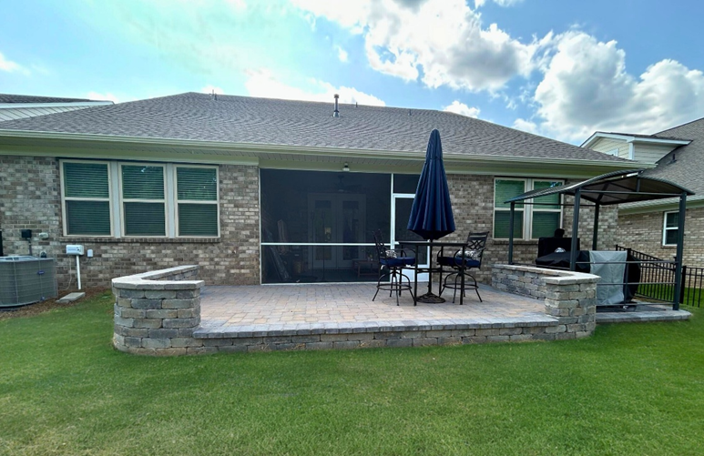 A spacious elevated stone paver patio with a curved retaining wall, a dining table with an umbrella, and a separate grilling station under a small gazebo, attached to a brick home with a screened porch, in Mint Hill, NC.