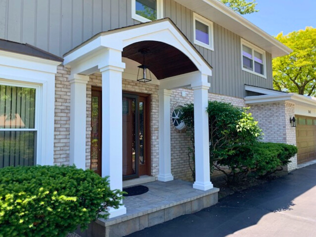A newly built covered front porch with white columns and a dark wood ceiling, complementing a house with grey siding and brick accents.