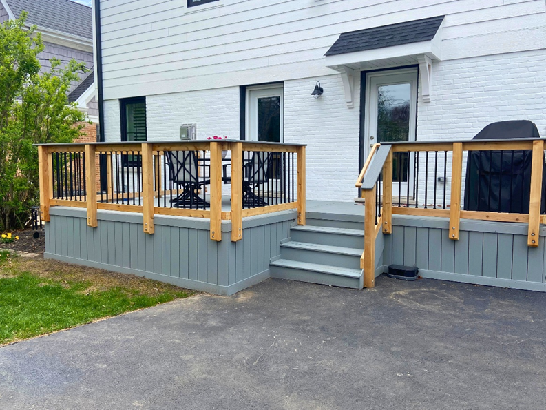 A ground-level deck with a gray-painted fascia and two matching gray steps leading up to it. The deck's railing system is made of natural, unstained wood posts and a top rail, with black metal balusters. A white brick wall with a door is in the background.