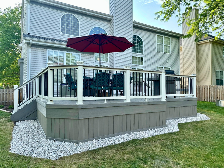 : A custom-built, multi-level composite deck with grey planks and white railings, featuring a dining area with a red umbrella and a grilling station, surrounded by white decorative stones and green lawn, attached to a light-colored home in Geneva, IL.