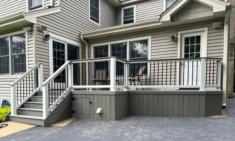 A newly built gray deck with a dark top railing and white balusters. A set of steps leads down from the deck. A small door for built-in storage is visible under the deck platform. The side of the house is covered in light gray siding.