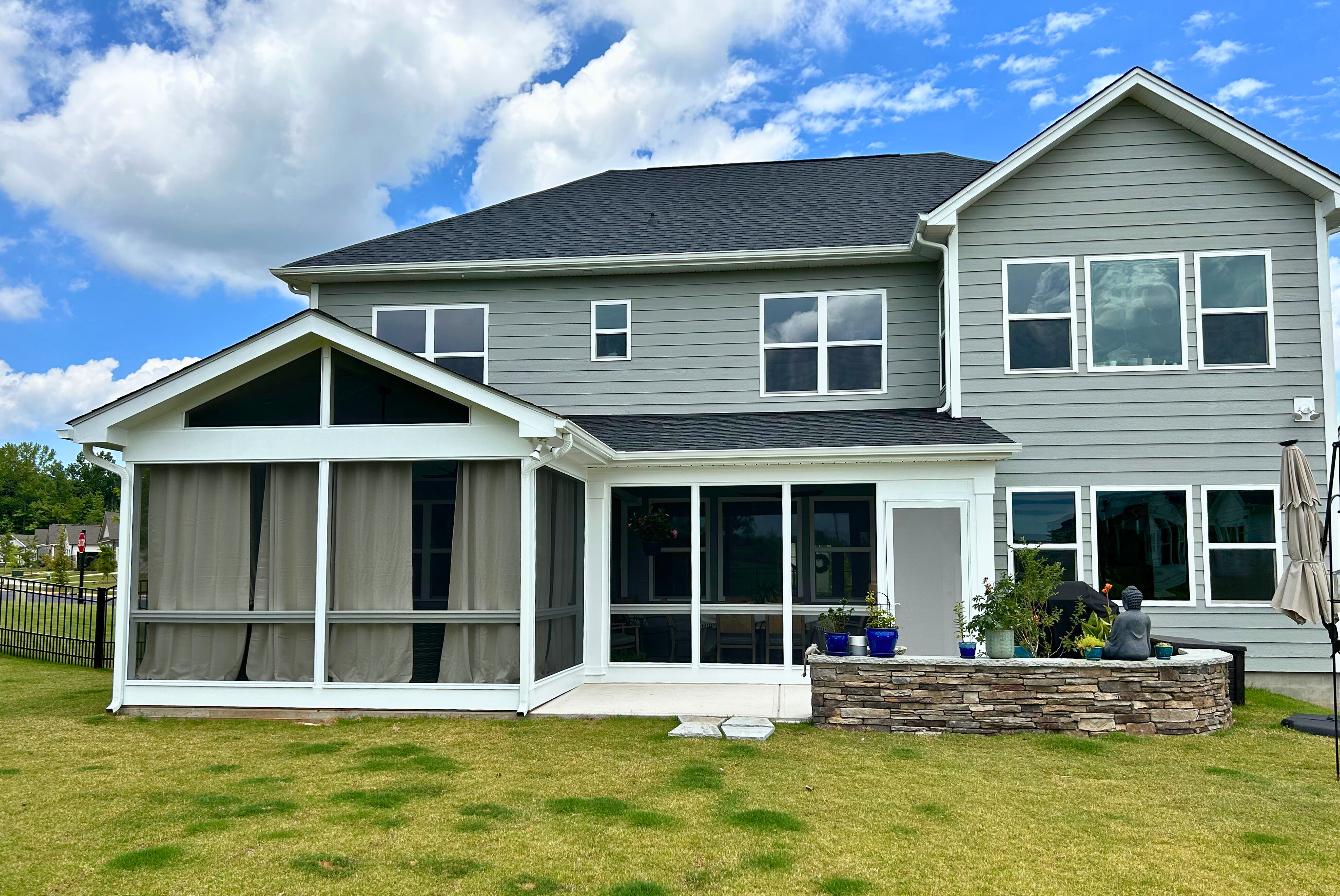 A modern gray house with a newly constructed screened porch featuring large windows and a stone accent wall, leading to a small patio area with a stone bench.