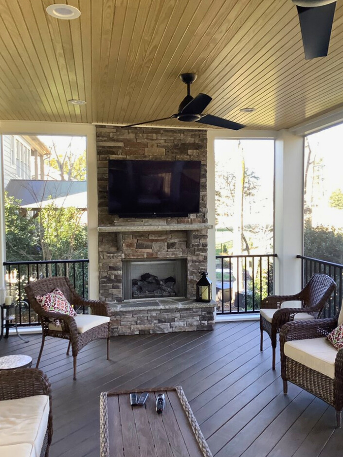 Interior view of a luxurious covered porch featuring a stone fireplace with a mounted television, comfortable wicker seating, and a wood plank ceiling with a ceiling fan.