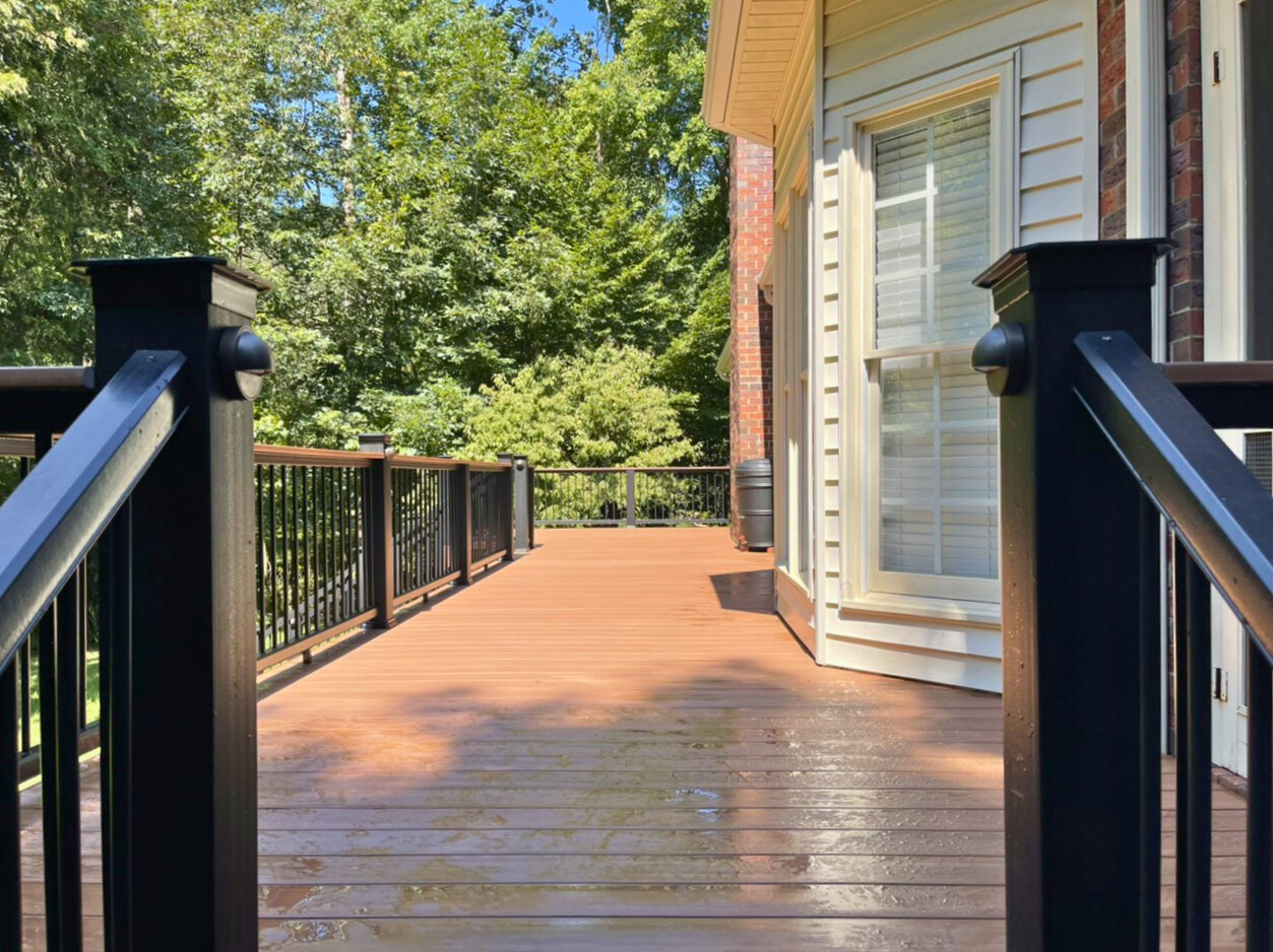 A spacious, modern composite deck with dark brown planks and black railings, extending from a house with white siding and brick accents, surrounded by green trees.