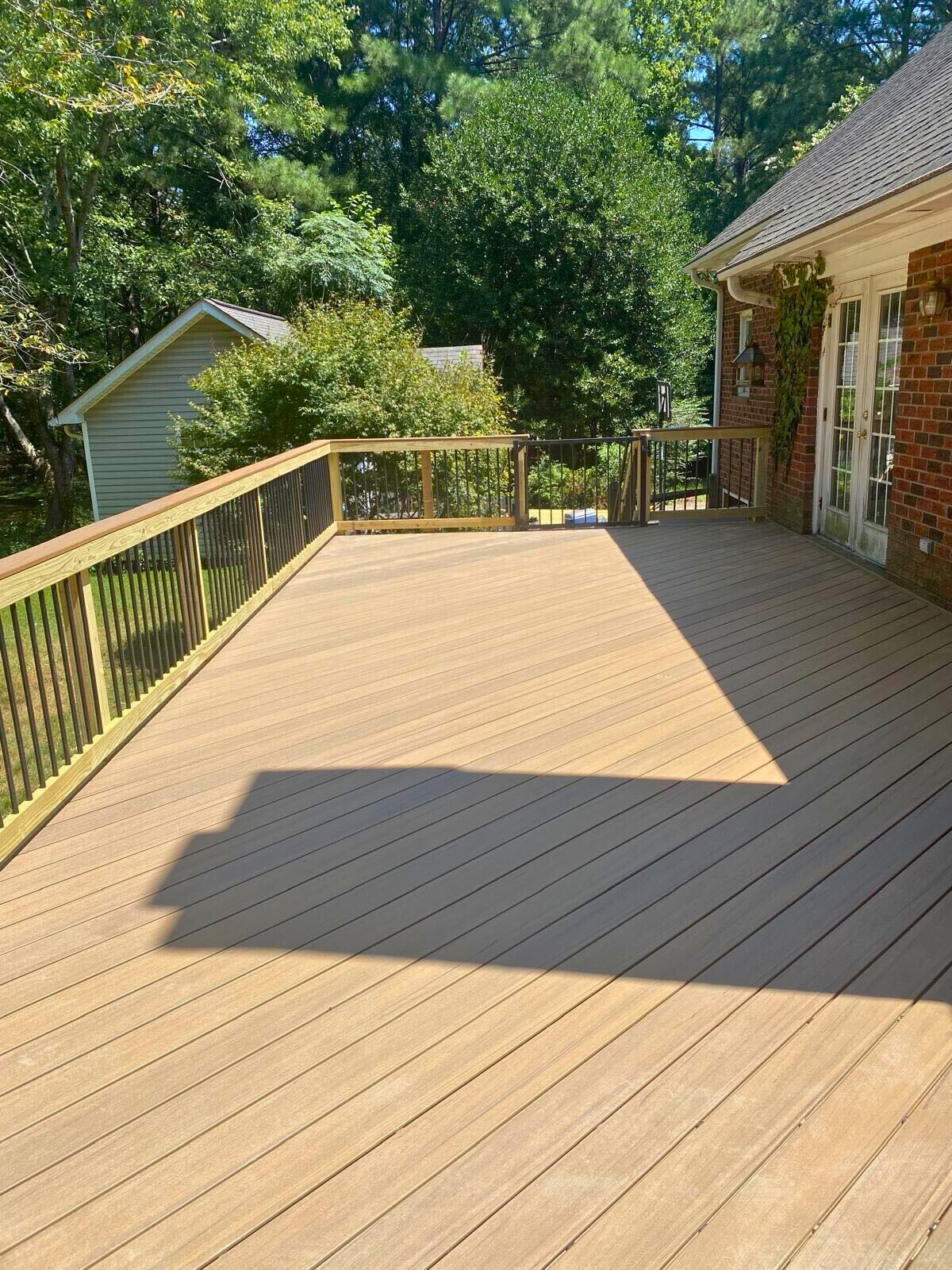 Large, new Trex Havana Gold composite deck with black metal railings, extending from a brick house in Matthews, NC, with trees in the background.