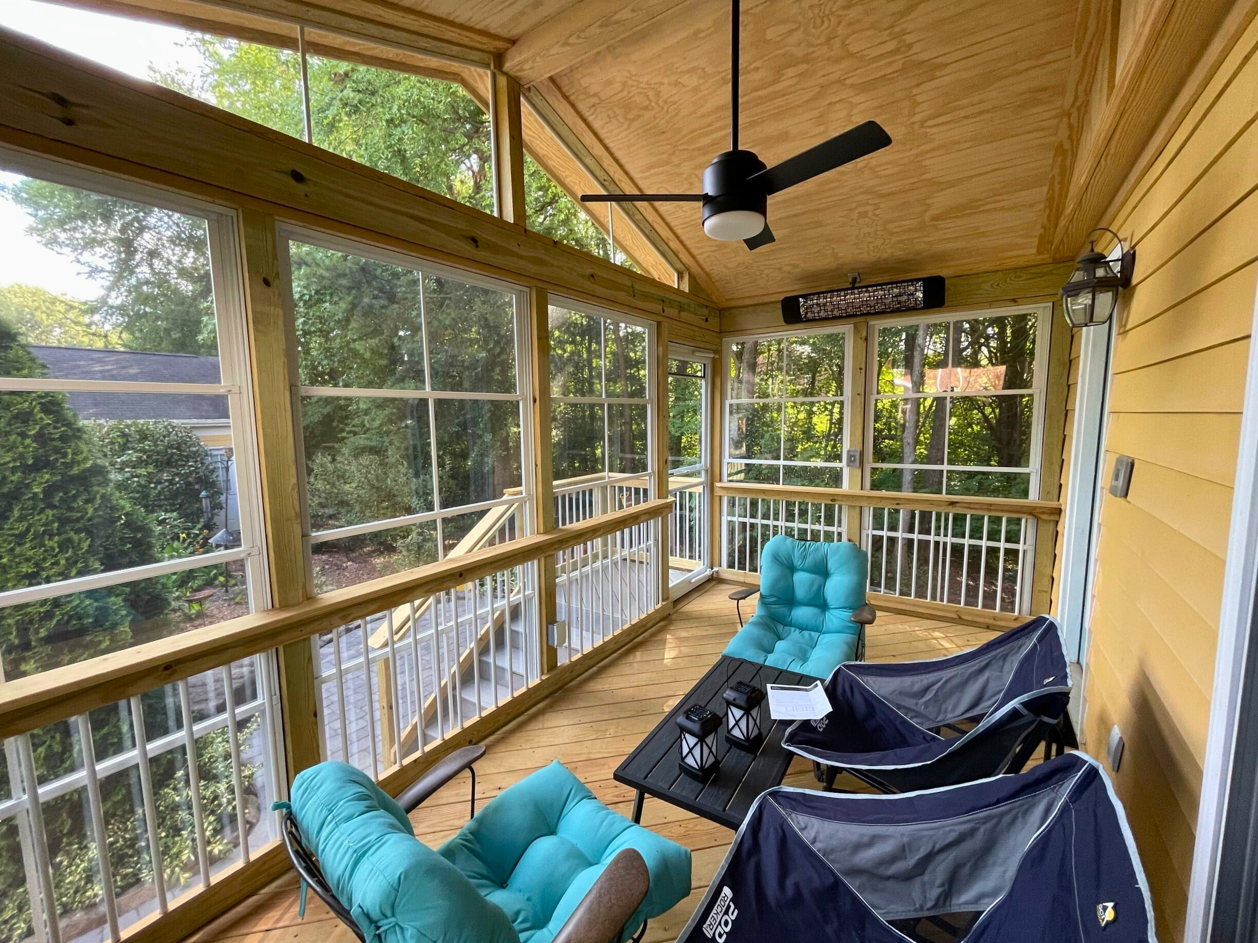 Interior of a bright Eze-Breeze porch with large vertical windows, light wood paneling on the walls and ceiling, comfortable seating, and a ceiling fan, overlooking a treed backyard.