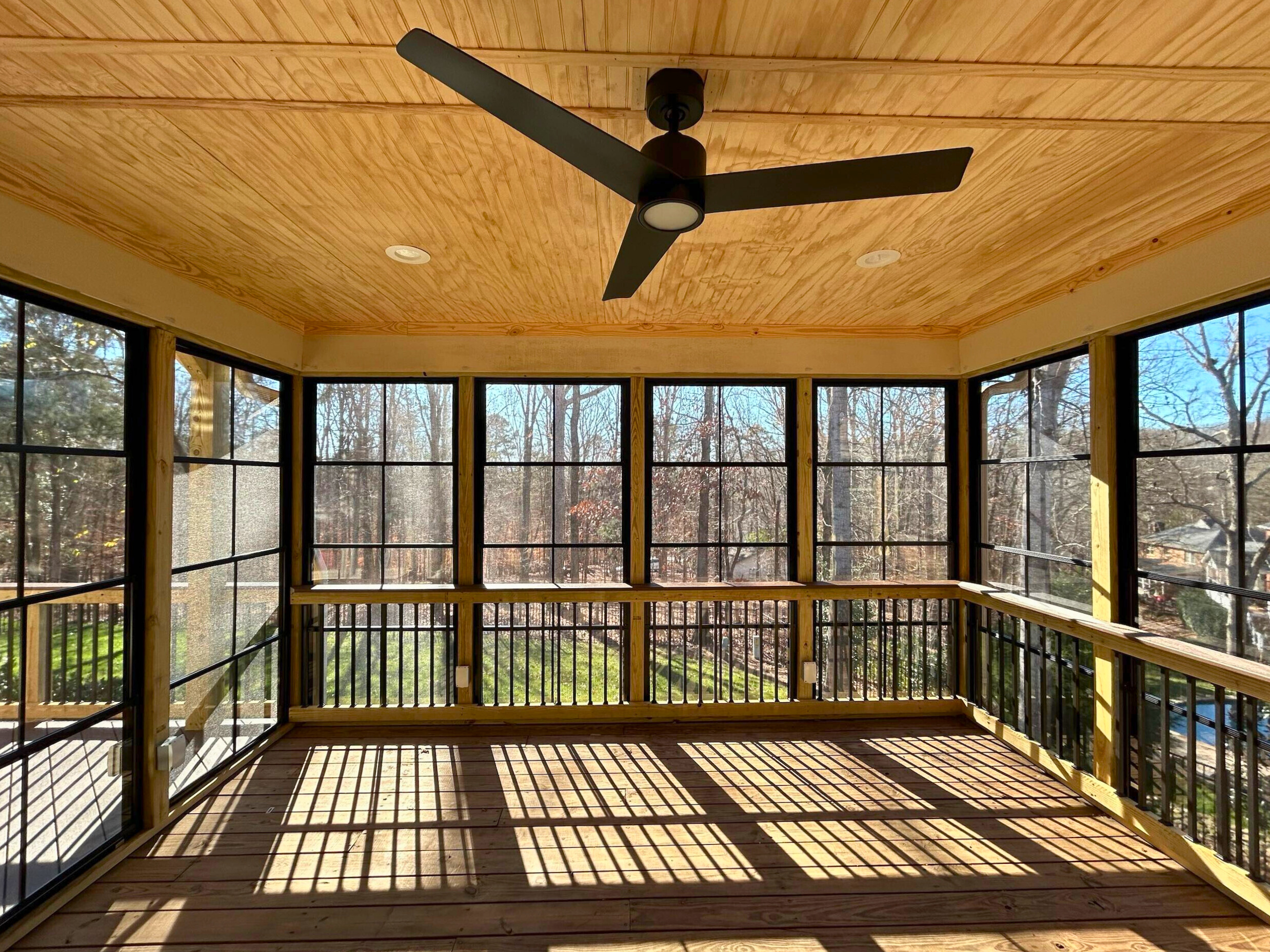 Interior view of a bright screened porch in Matthews, NC, with a wooden ceiling, ceiling fan, and large windows looking out to trees.
