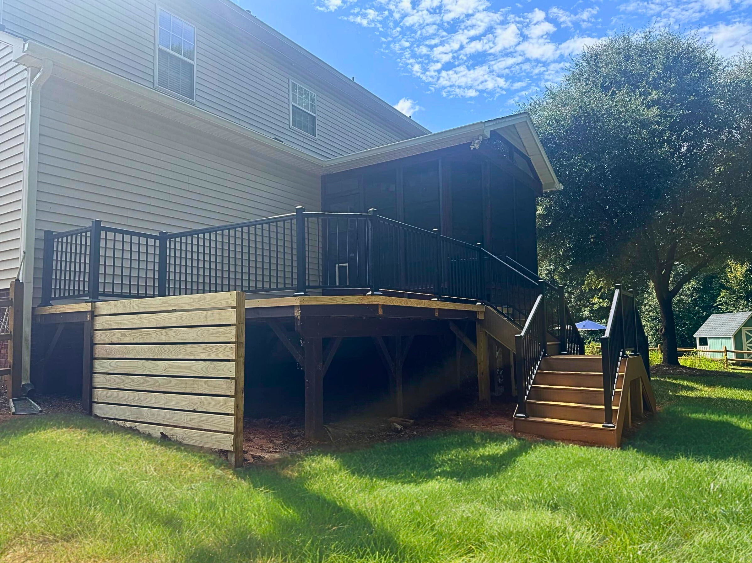 Modern composite deck with black metal railings and stairs leading down to a paver patio with a stone outdoor fireplace, at a brick house in Matthews, NC.