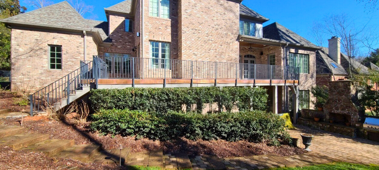 Modern composite deck with black metal railings and stairs leading down to a paver patio with a stone outdoor fireplace, at a brick house in Matthews, NC.