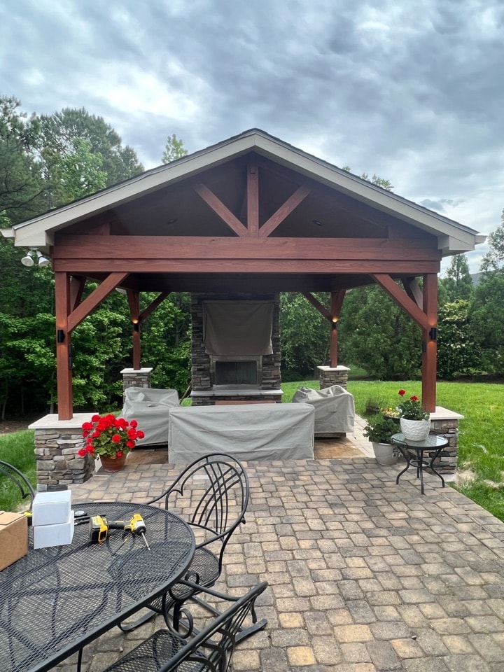 Custom outdoor pavilion in Matthews, NC, featuring a brown gable roof, stone fireplace, and stone columns, built on a paver patio with outdoor furniture.