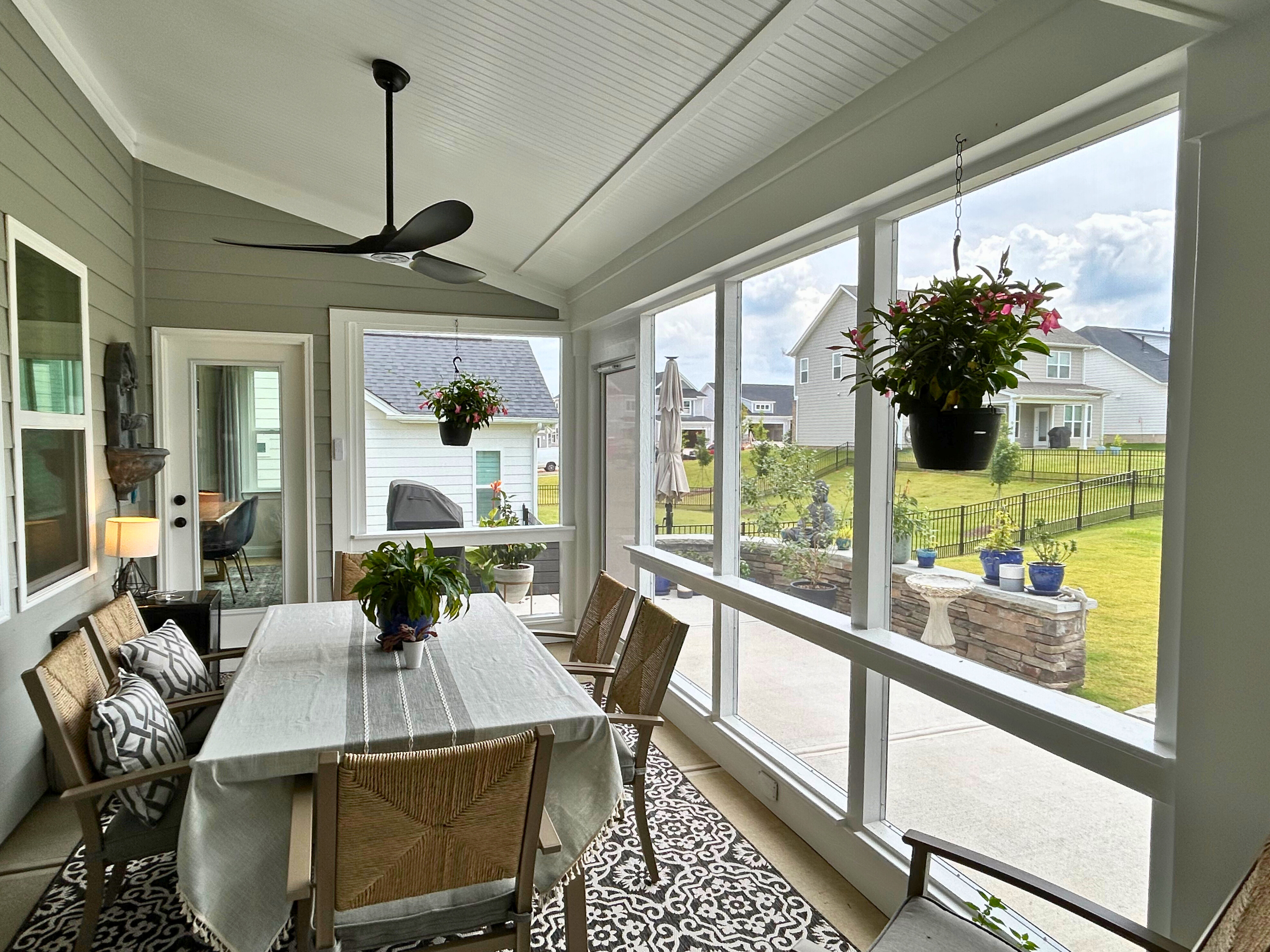 terior view of a spacious screened porch with a dining table and chairs, ceiling fan, and large windows looking out onto a green backyard with other houses in the distance.