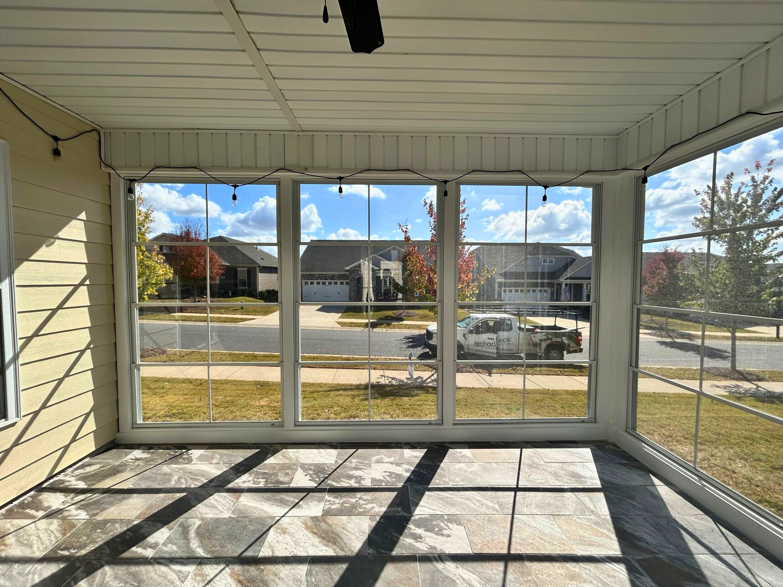 Interior of a three-season room with large windows, tiled flooring, string lights, and a view of a suburban neighborhood.