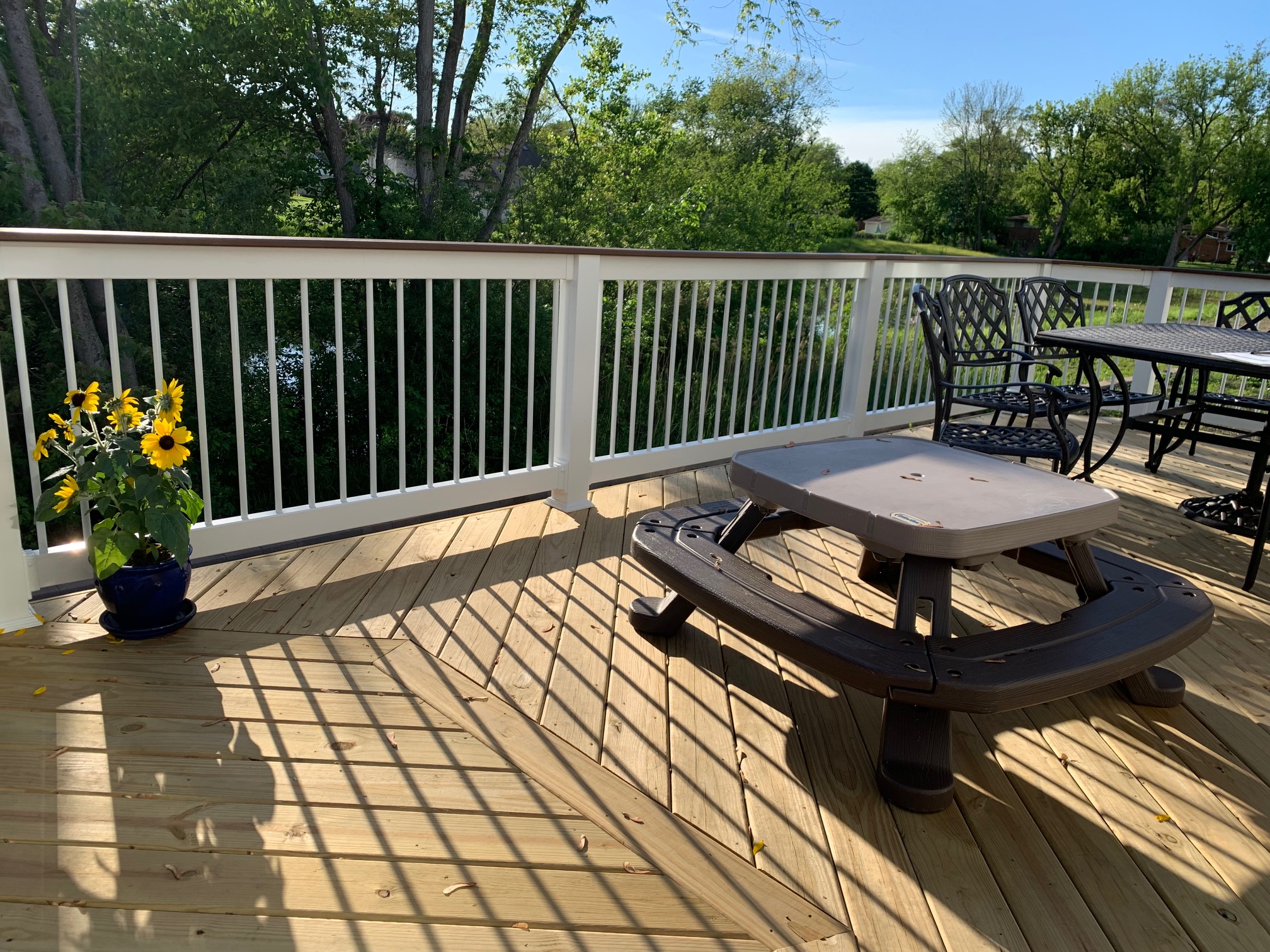 A newly built wooden deck with white railings and black balusters, featuring a picnic table and a potted sunflower, overlooking a lush green landscape.