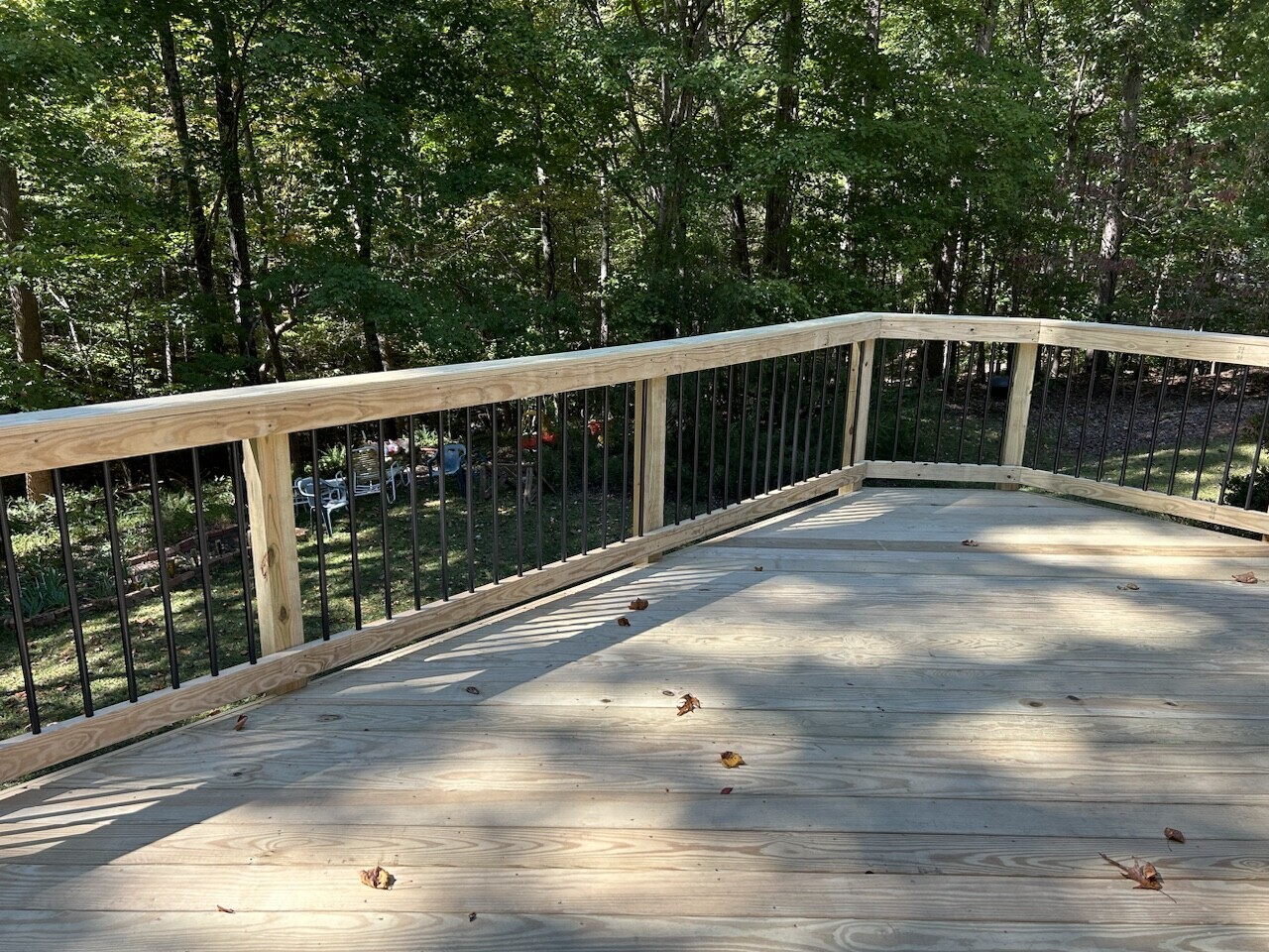 A newly built wooden deck with natural wood planks and a light wood railing with black balusters, overlooking a serene wooded backyard.